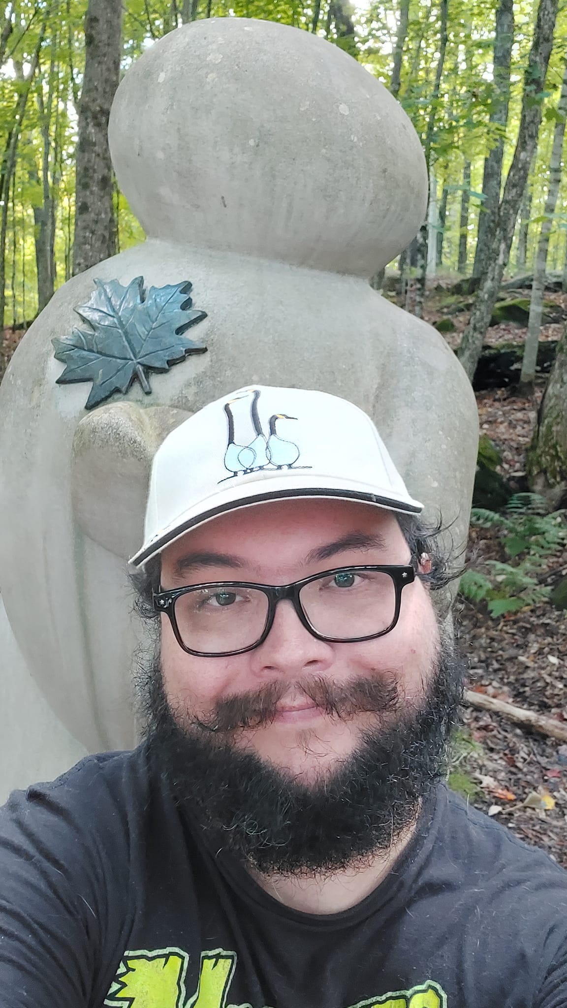 A man with short hair and bushy facial hair stands in front of a concrete structure shaped like a large person made of balloons, there is a green coroded copper casting of a maple leaf on its upper left chest